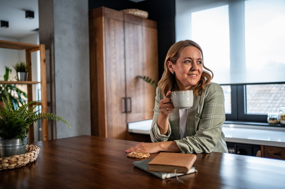 Happy mid adult woman enjoying a relaxing coffee break, smiling and looking away while sitting at a kitchen island, contemplating and planning her day