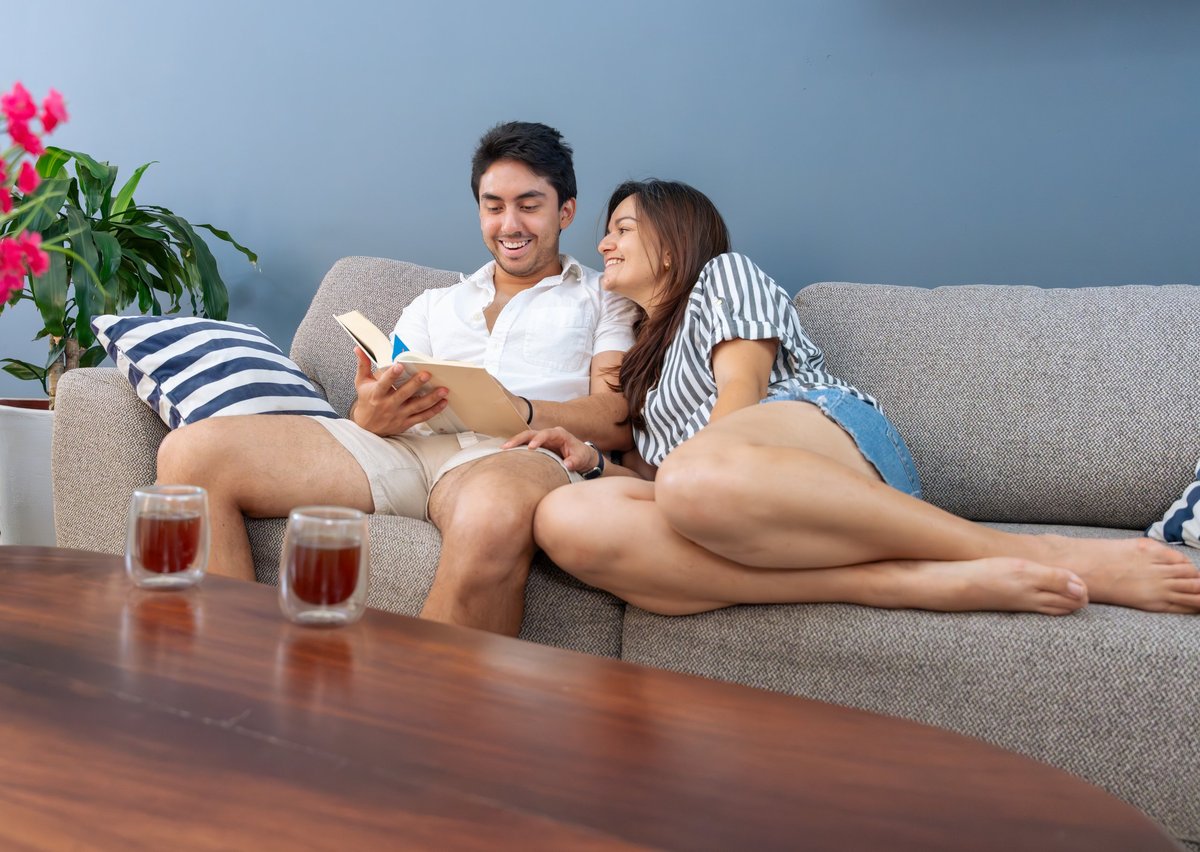 Young adult couple enjoying a quiet afternoon at home, relaxing on the sofa, with the man reading a book while the woman smiles, emphasizing cozy leisure and togetherness