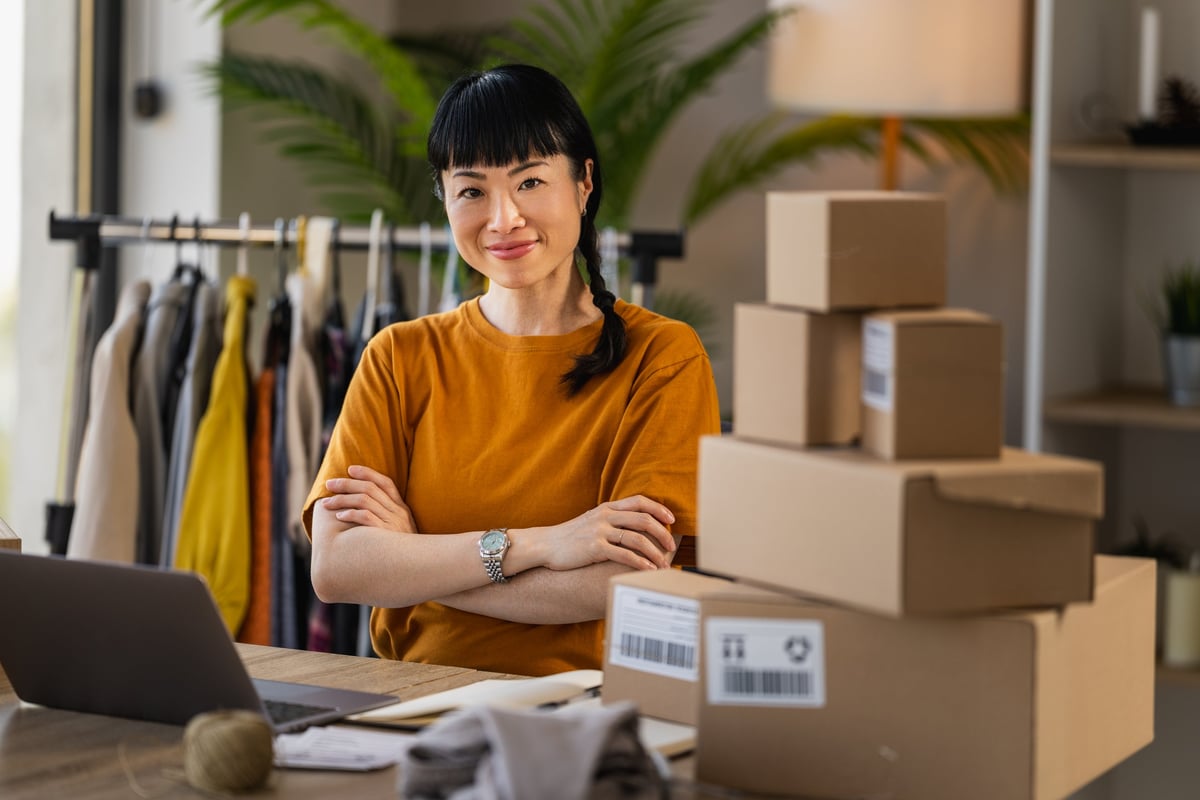 Small business owner standing confidently in a home workspace surrounded by packed shipping boxes, clothing rack and laptop, preparing orders for an online shop